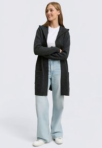 Temno siva jopica s kapuco, dolgimi rokavi in dvema sprednjima žepoma, kombinirana s svetlo modrimi širokimi kavbojkami in belimi supergami.