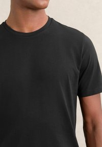Tricou negru din bumbac cu guler rotund, mâneci scurte și textură netedă; imaginea se concentrează pe partea superioară a toracelui și zona umărului.