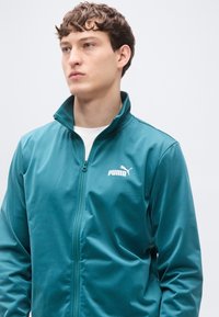 Τζάκετ με φερμουάρ σε τιρκουάζ χρώμα, κατασκευασμένο από λεία, ελαφριά ύλη, με ψηλό γιακά και λευκό λογότυπο Puma στο αριστερό στήθος.