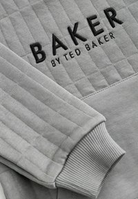 Šedá prešívaná mikina s vykrajovaným textom "BAKER BY TED BAKER." Rúbkované manžety s hladkou textúrou, kontrastné šitie a obdĺžnikové vzory.