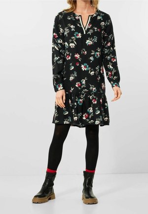 Femme portant une robe longue à manches longues noire à motifs floraux, des collants noirs, des chaussettes rouges et des bottines noires à semelles marron, debout devant un fond uni.