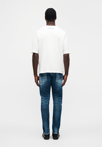 Λευκό κοντομάνικο T-shirt με στρογγυλή λαιμόκοψη και λογότυπο "DSQUARED2" στην πλάτη, σε συνδυασμό με μπλε distressed τζιν παντελόνι.