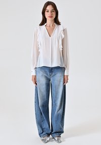 Blusa bianca e trasparente con dettagli arricciati, scollatura a V e maniche lunghe, abbinata a jeans di denim azzurro chiaro oversize. Scarpe argentate a punta.