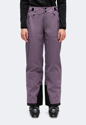 Modèle portant un pantalon de ski ample violet avec des poches zippées et des chaussures de ski noires, sur un fond uni.