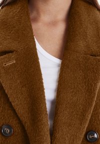 Brun, struktureret uldfrakke med dobbeltradet design, store sorte knapper og en bred krave, lagdelt over en hvid top.