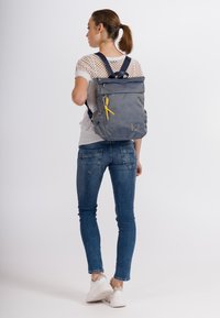Mochila gris con diseño de tapa enrollable, que presenta un acento amarillo, correas ajustables y una forma estructurada, mostrada en una persona.