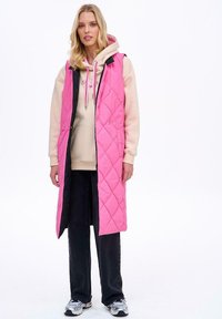 Różowa pikowana długa kamizelka z czarnymi akcentami, z kapturem, zatrzaskami i bocznymi kieszeniami, noszona na jasnym beżowym hoodie i czarnych spodniach.