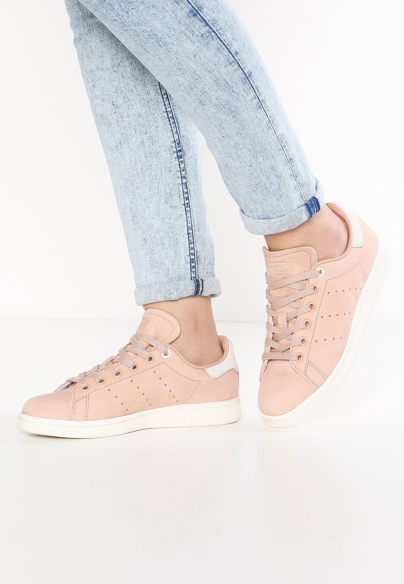 Fötter i ljusrosa mockaskor med vita sulor och ljusblå upprullade jeans framför en enkel vit bakgrund.