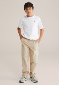 Balta kokvilnas t-krekls ar mazu melnu logotipu, apvienots ar brīvām bēšām biksēm un pelēkiem sporta apaviem ar baltām akcentiem un melniem zoliem.