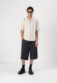 Stribet kortærmet skjorte i beige og creme, parret med mørkegrå knæ-lange shorts. Båret med sorte snørebåndsstøvler og solbriller.