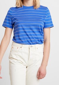 Niebieski t-shirt z krótkim rękawem w białe poziome paski, zestawiony z jasnymi, przetartymi dżinsami. Prosty, okrągły dekolt.