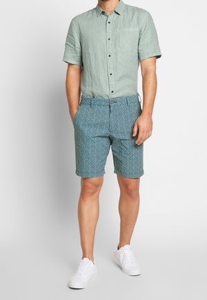 Leinen-Kurzarmhemd in Hellgrün, kombiniert mit blauen, gemusterten Shorts. Weiße Sneakers vervollständigen das Outfit. Sichtbare Knöpfe und Taschen.
