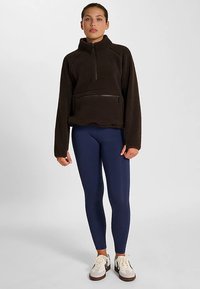 Brauner Fleece-Pullover mit hohem Kragen, halbseitigem Reißverschluss und Fronttasche. Kombiniert mit dunkelblauen Leggings und beigen Sneakers. Einfaches Design.