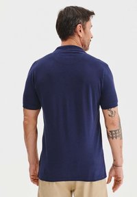 Granatowa koszulka polo z krótkim rękawem, o strukturze materiału i ściągaczem przy kołnierzyku. Posiada gładkie plecy bez widocznych znaków towarowych czy wzorów.