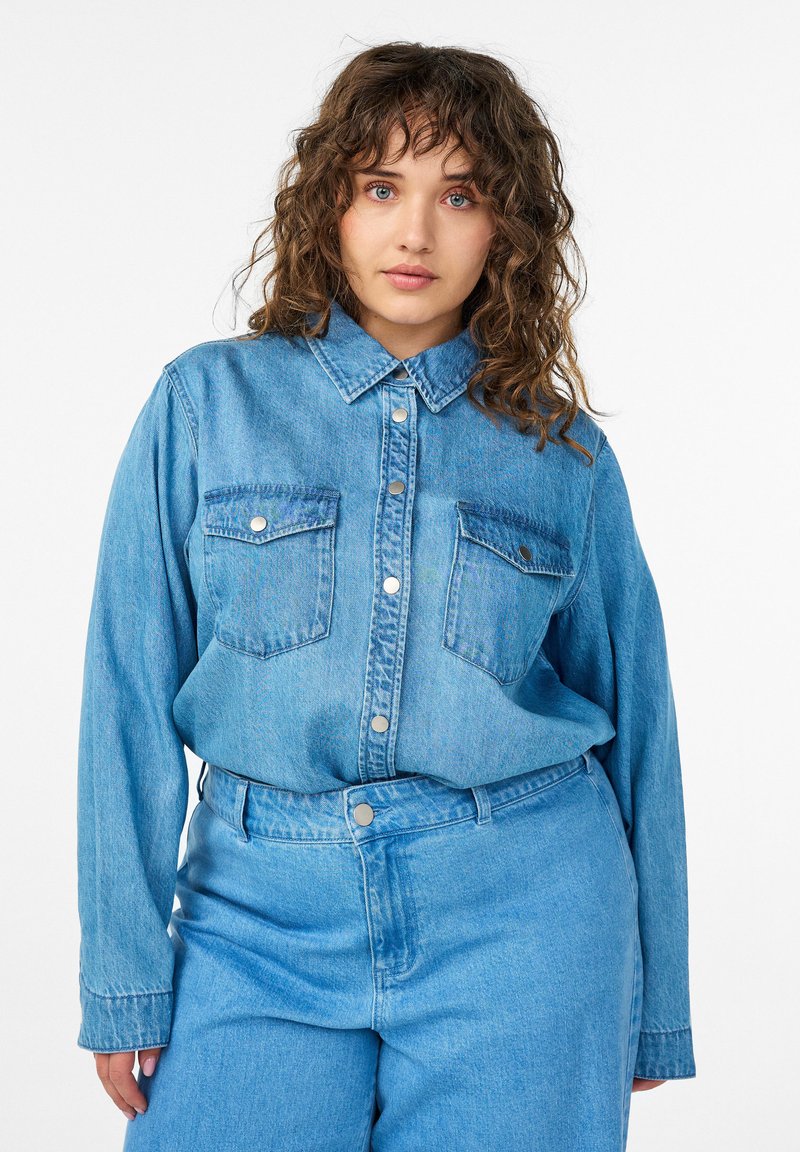 Žena s kučeravými hnedými vlasmi, oblečená v modrej džínsovej košeli a zodpovedajúcich rifle s vysokým pásom, stojaca na bielom pozadí, hľadiaca dopredu.