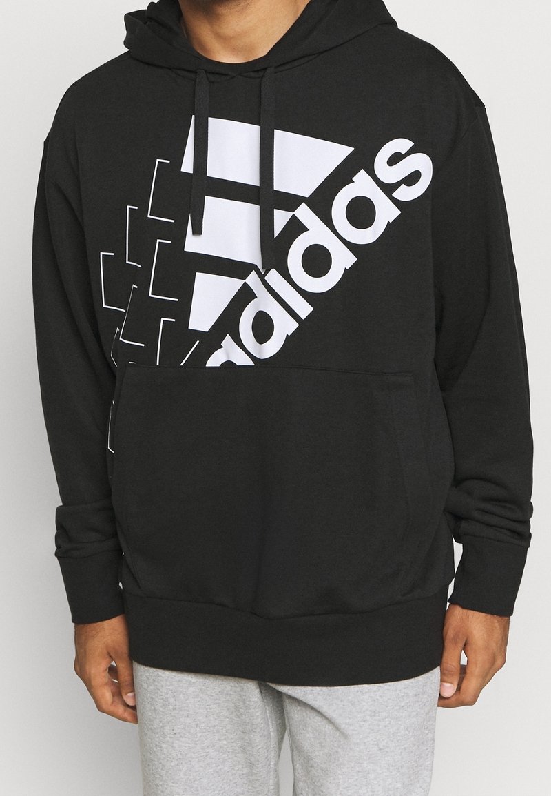 Homme portant un sweat à capuche noir Adidas avec un grand logo blanc et un pantalon de survêtement gris, debout devant un fond uni.