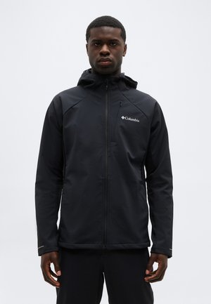 Άνδρας που φοράει μια μαύρη κουκούλα μπουφάν Columbia με φερμουάρ κλειστό, στέκεται μπροστά σε απλό ανοιχτό φόντο, κοιτάζοντας προς τα εμπρός.