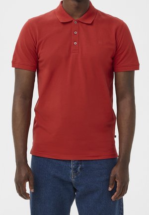 Mann trägt ein rotes Polohemd mit kurzen Ärmeln und blaue Jeans vor einfarbigem Hintergrund, beschnitten am Hals und in der Mitte des Oberschenkels.