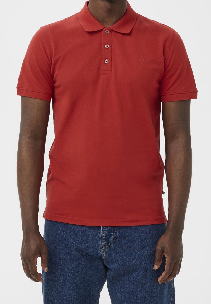 Homme portant un polo rouge à manches courtes et un jean bleu en denim sur fond uni, cadré au niveau du cou et du milieu de la cuisse.