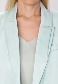 Femme portant un blazer bleu pastel clair par-dessus un haut à col en V assorti, mettant en valeur le décolleté et la partie supérieure de la poitrine.