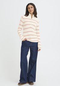 Jersey de punto en crema con rayas horizontales rosas, cuello con cremallera y mangas largas. Combinado con jeans de pierna ancha en azul oscuro y zapatos marrones.