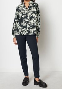 Chemise à imprimé floral vert foncé avec des fleurs blanches, dotée d'un col et de manches longues. Associée à un pantalon bleu marine foncé et des mocassins compensés noirs.