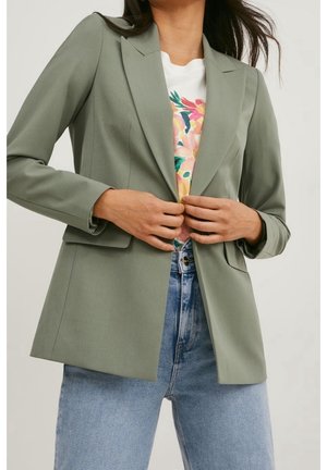 Mujer abrochándose una blazer verde claro sobre una camisa blanca con estampado floral, vistiendo vaqueros de tiro alto azul claro contra un fondo liso.