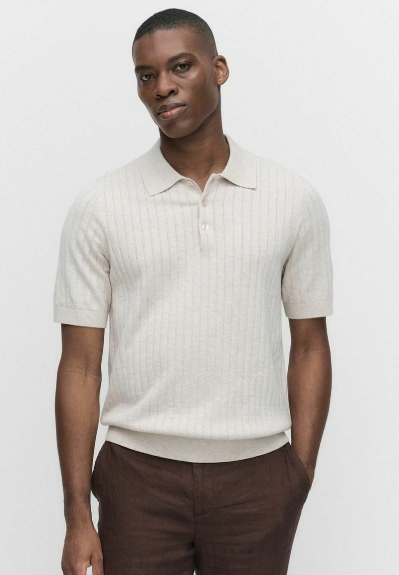 Kortärmad, ljusbeige polo skjorta i ribbat tyg; har en krage och en rak fåll. Bärs med bruna byxor.