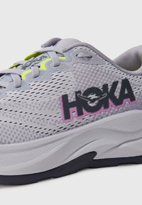 Szara sportowa but z siateczkową cholewką, akcentowana jasnoszergeniowymi sznurówkami oraz wyraźnym czarnym i różowym logo "HOKA". Wyściółka w podeszwie.