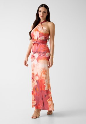 Femme aux cheveux longs et foncés portant une robe longue jusqu'au sol, à col licou, fleurie dans des tons orange, rose et crème, se tenant avec un bras détendu.