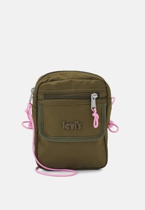 Bolso bandolera de tela verde oliva con correas de acento rosa, bolsillo frontal con cremallera y logotipo en relieve en la parte delantera. Forma rectangular y compacta.