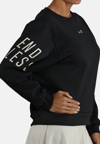 Sudadera negra de algodón, con mangas largas. La manga izquierda tiene texto blanco que dice "ENDLESS" y un pequeño logo en el pecho.