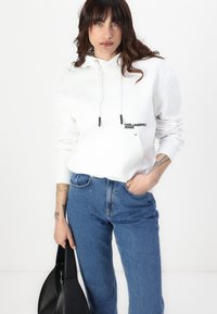 Sudadera blanca con logo negro "KARL LAGERFELD JEANS", cordones y bolsillo canguro, combinada con jeans azules y un bolso negro.