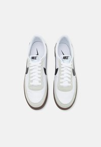 Zapatillas Nike con parte superior de cuero blanco, detalles en gamuza gris, logotipo Swoosh negro, cordones blancos y suela de goma marrón clara.