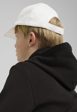 Gorra de béisbol blanca con visera curva, usada al revés, combinada con un sudadero negro con capucha. El sudadero tiene una textura en la tela.