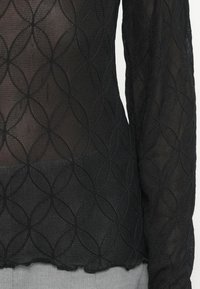 Bluză lungă neagră cu mâneci lungi, realizată dintr-un material transparent cu un model geometric de cercuri suprapuse. Marginea de jos este ușor desfăcută.
