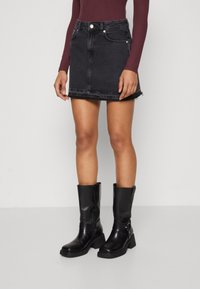 Ben som bär en svart denim mini-kjol med fransad fåll, svarta läderstövlar som når midjan och en långärmad vinröd topp mot en enkel bakgrund.