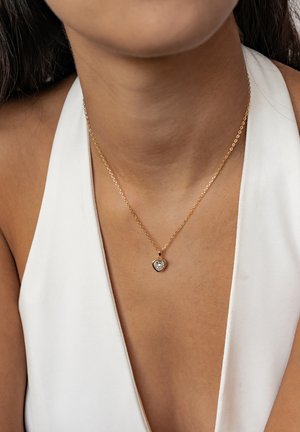 Gold chain necklace featuring a heart-shaped pendant with a clear gemstone. The pendant is outlined in gold, resting on bare skin.