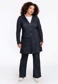 Donker spijkerjack met een opvallende kraag, vijf knopen en een gestructureerde vorm. Gepaard met bijpassende donkere spijkerbroek en witte schoenen.