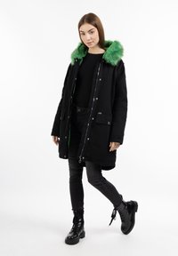 Parka nera con cappuccio in pelliccia sintetica verde, due tasche frontali e chiusura con zip. Indossata con jeans neri aderenti e stivali neri con lacci.