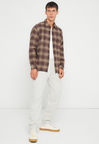 Brun rutete flanellskjorte med knapper, brukt over en hvit t-skjorte, kombinert med grå joggebukser og kremfargede joggesko.