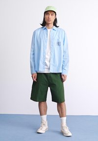 Camisa de botones a rayas azul claro con el logo "OBEY", camiseta blanca, pantalones cortos verde oscuro, gorra verde claro y deportivas beige con detalles blancos.