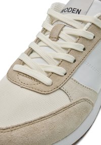 Zapatillas en beige y blanco que presentan materiales de ante y malla, cordones planos y un logo en la lengüeta. Costuras decorativas a lo largo de los bordes.
