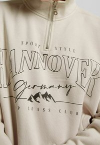 Ljusbeige tröja med kvartsyz, tillverkad av mjukt tyg. Har "HANNOVER" i feta, svarta bokstäver, med en bergsgrafik nedanför.