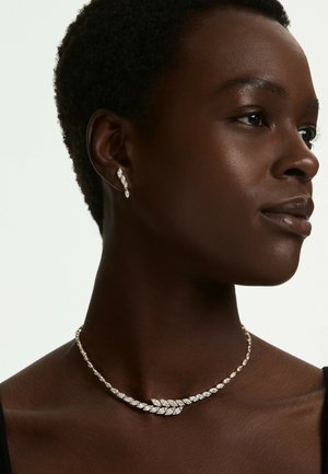 Vrouw met kort zwart haar die een fonkelende bladvormige diamanten ketting en bijpassende hangoorbellen draagt, naar rechts kijkend tegen een effen achtergrond.