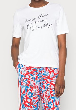 T-shirt en coton blanc à manches courtes avec un texte manuscrit noir. Associé à un pantalon rouge à motif floral avec des accents bleus et blancs.