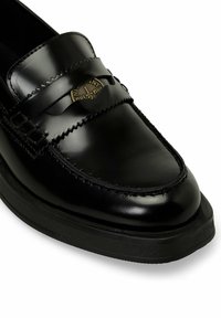 Mocassino in pelle nera con finitura liscia, caratterizzato da una fascia decorativa e un emblema in tonalità oro sulla parte superiore. Suola in gomma con texture bassa.