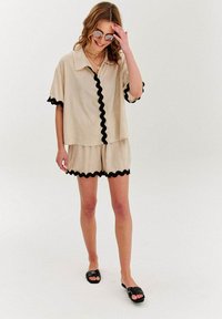 Beige košile s krátkým rukávem a černým rickrack lemováním a ladící šortky, které mají uvolněný střih. Model má na sobě černé šlapky a sluneční brýle.