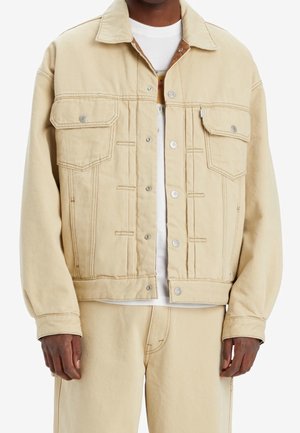 Mann trägt beige Jeansjacke und passende Hose, mit weißem Hemd darunter, steht mit entspannten Armen seitlich am Körper.