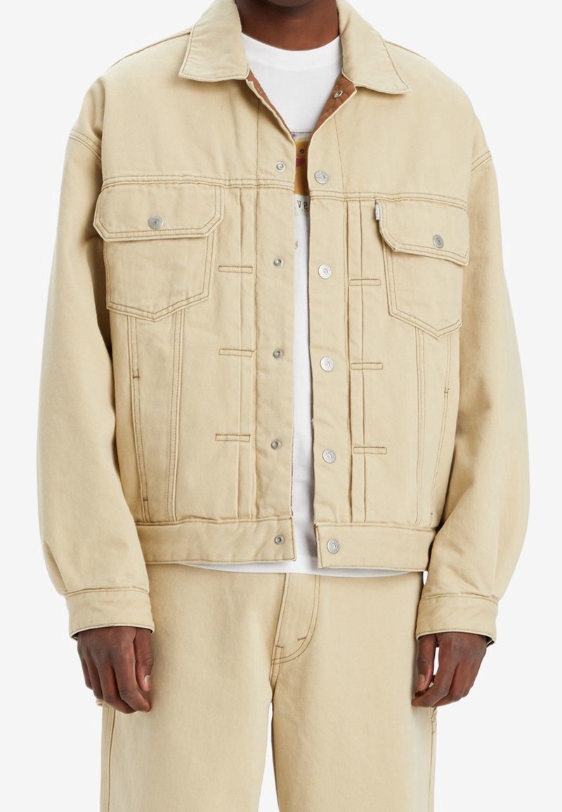 Homme portant une veste en denim beige et un pantalon assorti, avec une chemise blanche en dessous, debout les bras détendus le long du corps.